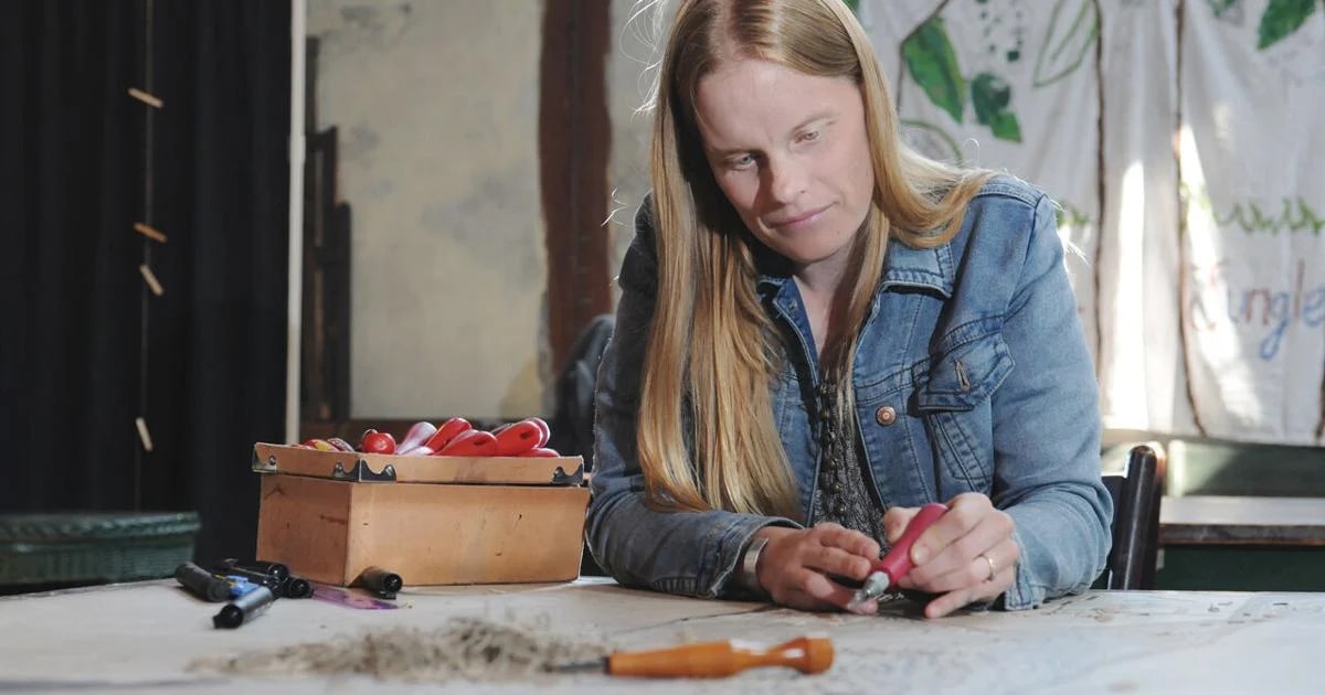 Christine Benson at a worktable carving with a red-handled tool, a box of carving tools beside her.