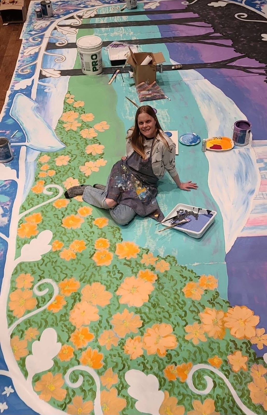A woman in a paint-splattered apron sitting on a large painted stage backdrop of orange flowers, with paint trays and brushes around her.