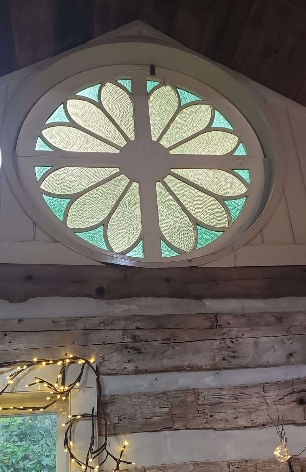 The round stained-glass window with green and pale yellow petals, high in the gable of the log-cabin studio.