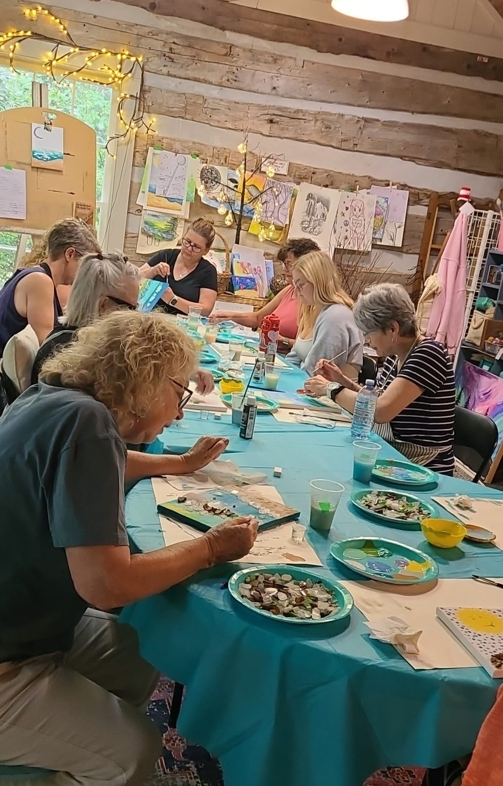 Adults at a long turquoise table in the log-cabin studio, painting small canvases and arranging pieces of sea glass.