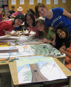 Five smiling kids leaning over a table to show off their finished landscape paintings.