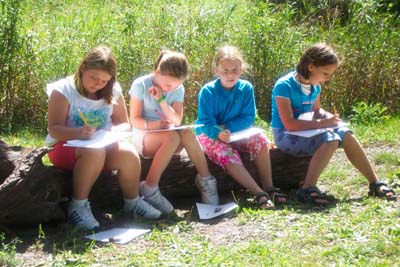 Four girls sitting on a fallen log outdoors, sketching on paper in the sun.