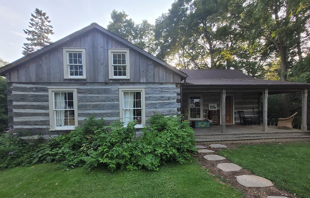 The Journey Through the Arts studio - a two-storey weathered log cabin with white-trimmed windows and a covered porch, set among trees in Port Hope.
