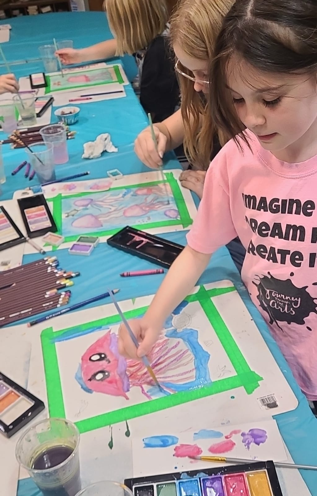 A girl in a Journey Through the Arts T-shirt painting a watercolour jellyfish, with other kids painting at the table beside her.