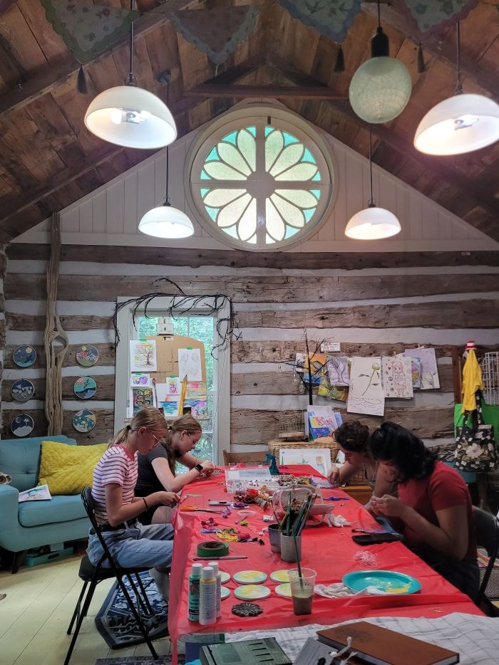 Older kids working on craft projects at a long red table in the log-cabin studio, under the round stained-glass window.