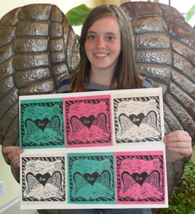 A girl holding up a sheet of six lino-cut prints of a winged heart in different colours.
