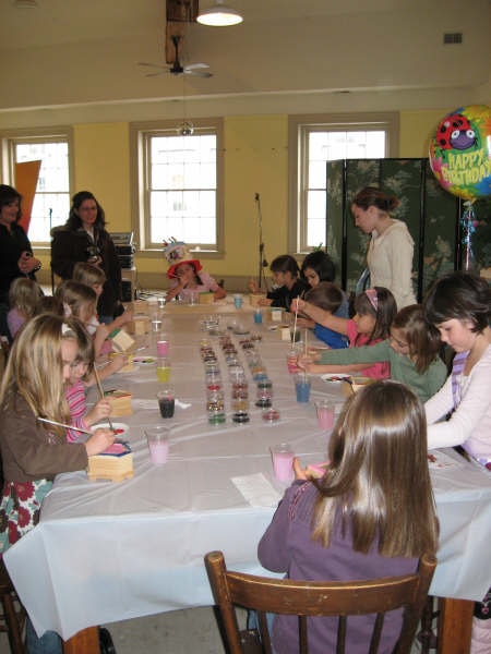Children painting small wooden boxes along a long table at a birthday party, with a Happy Birthday balloon in the corner.