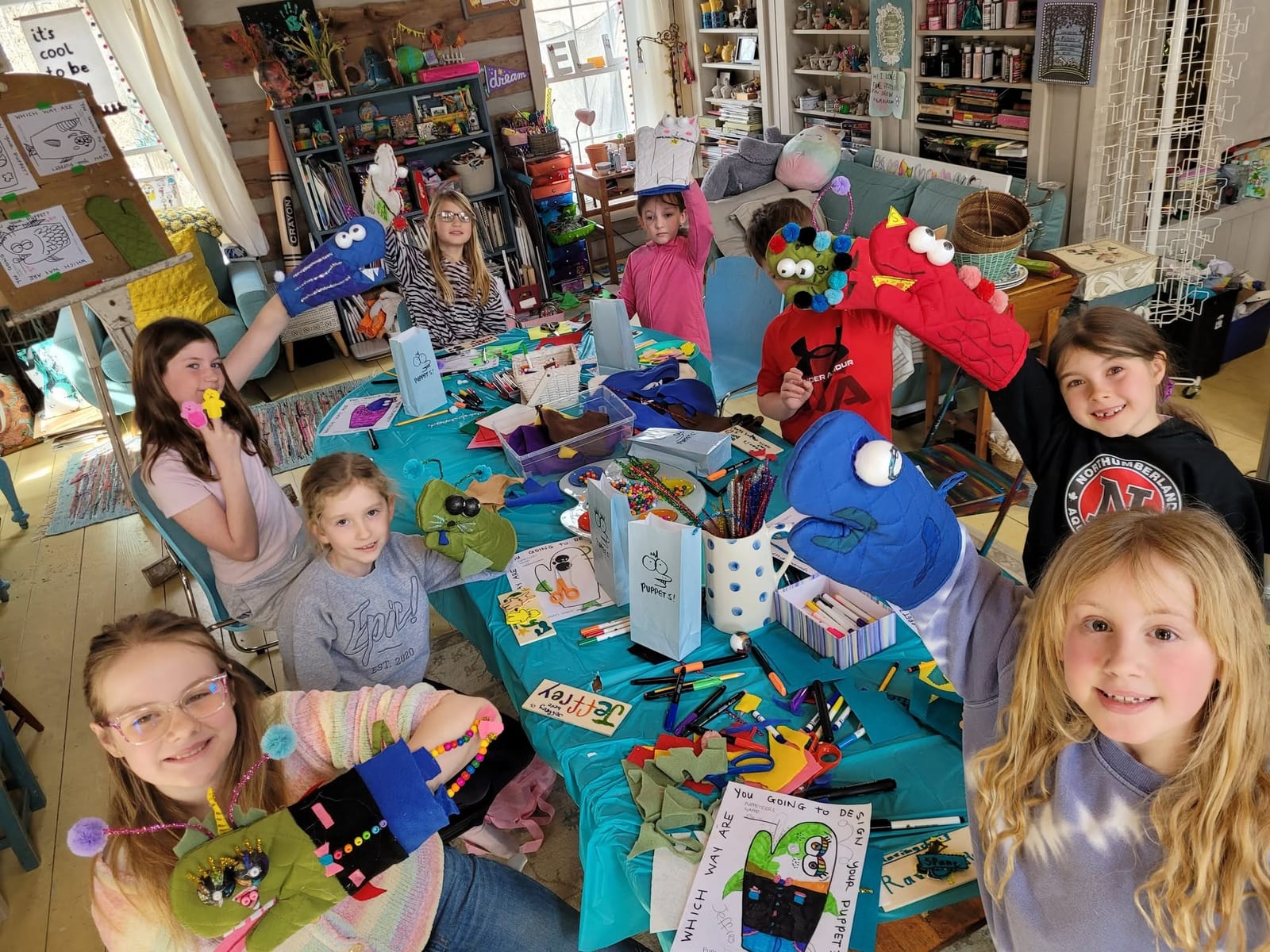 Children around a crowded craft table holding up the hand puppets they made.