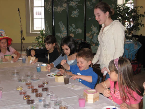 Children painting small wooden boxes at a table of glitter jars and paint cups while a young woman helps.