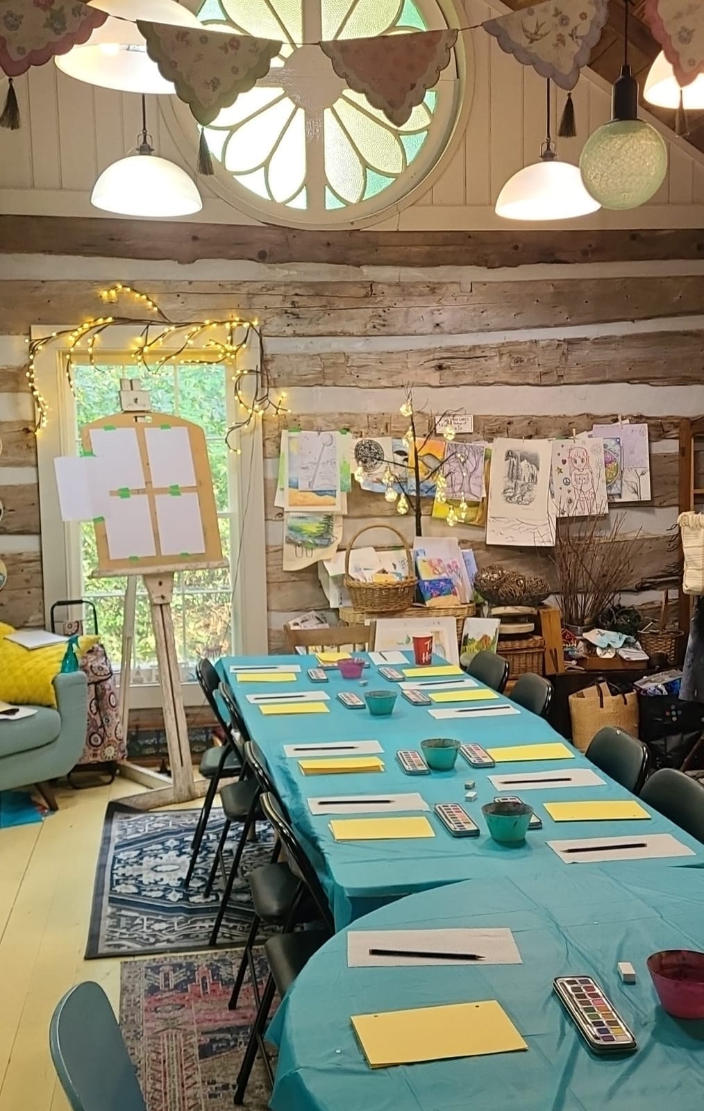 The log-cabin studio set up for a class, with paint sets and paper along a long table and children's art on the log walls.