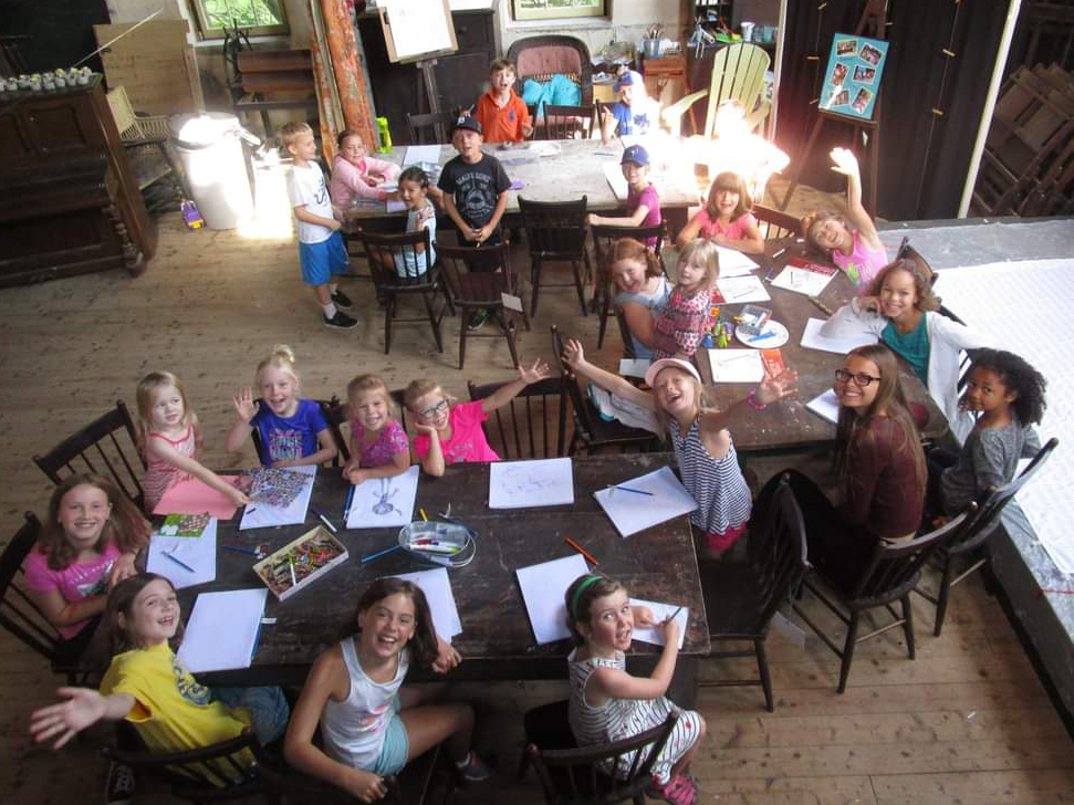 About twenty children seated around long wooden tables in the rustic cabin studio, drawing and waving at the camera.