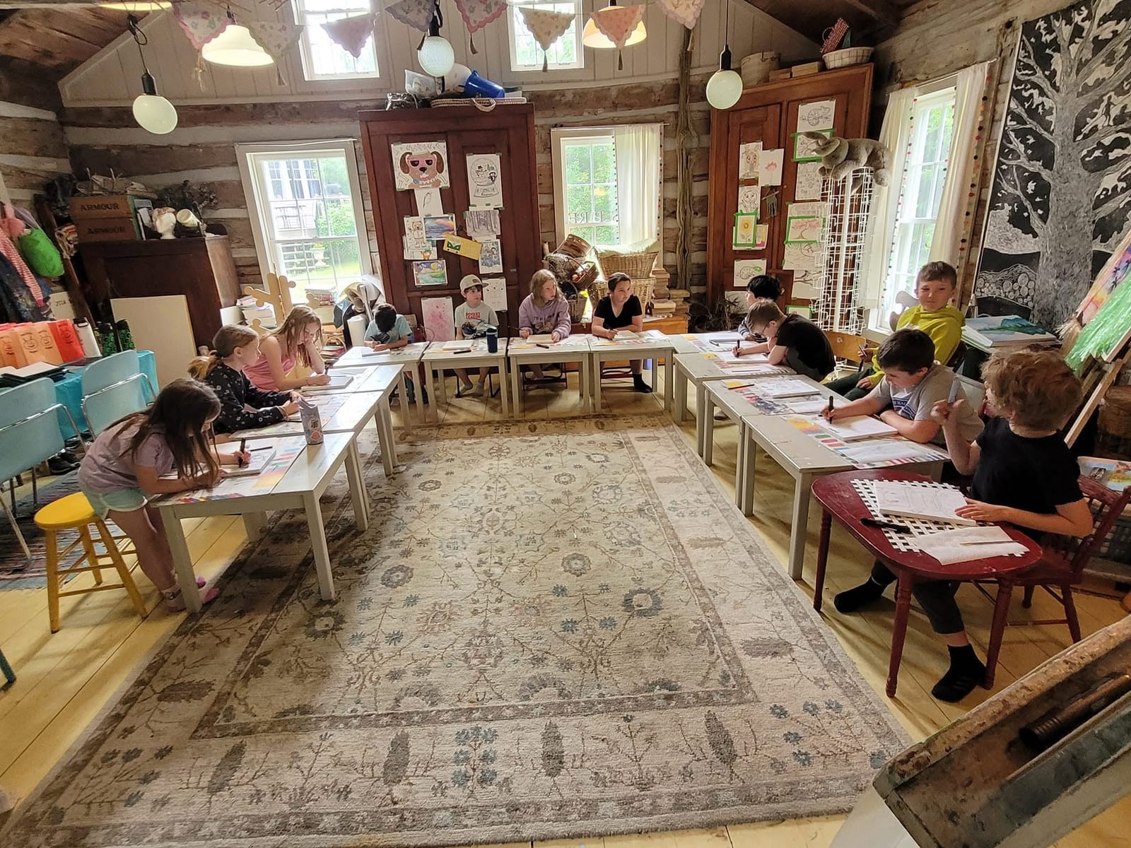 Children drawing at long tables set around a rug in the log-cabin studio.