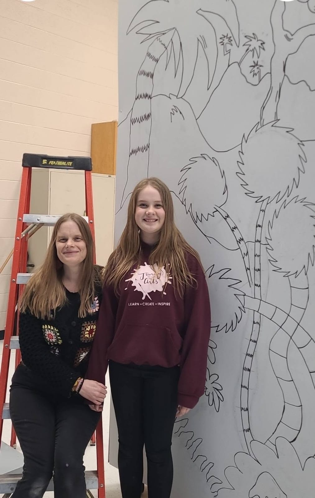 An adult and a teen in a Journey Through the Arts hoodie standing in front of a large mural outline drawn on a wall panel.