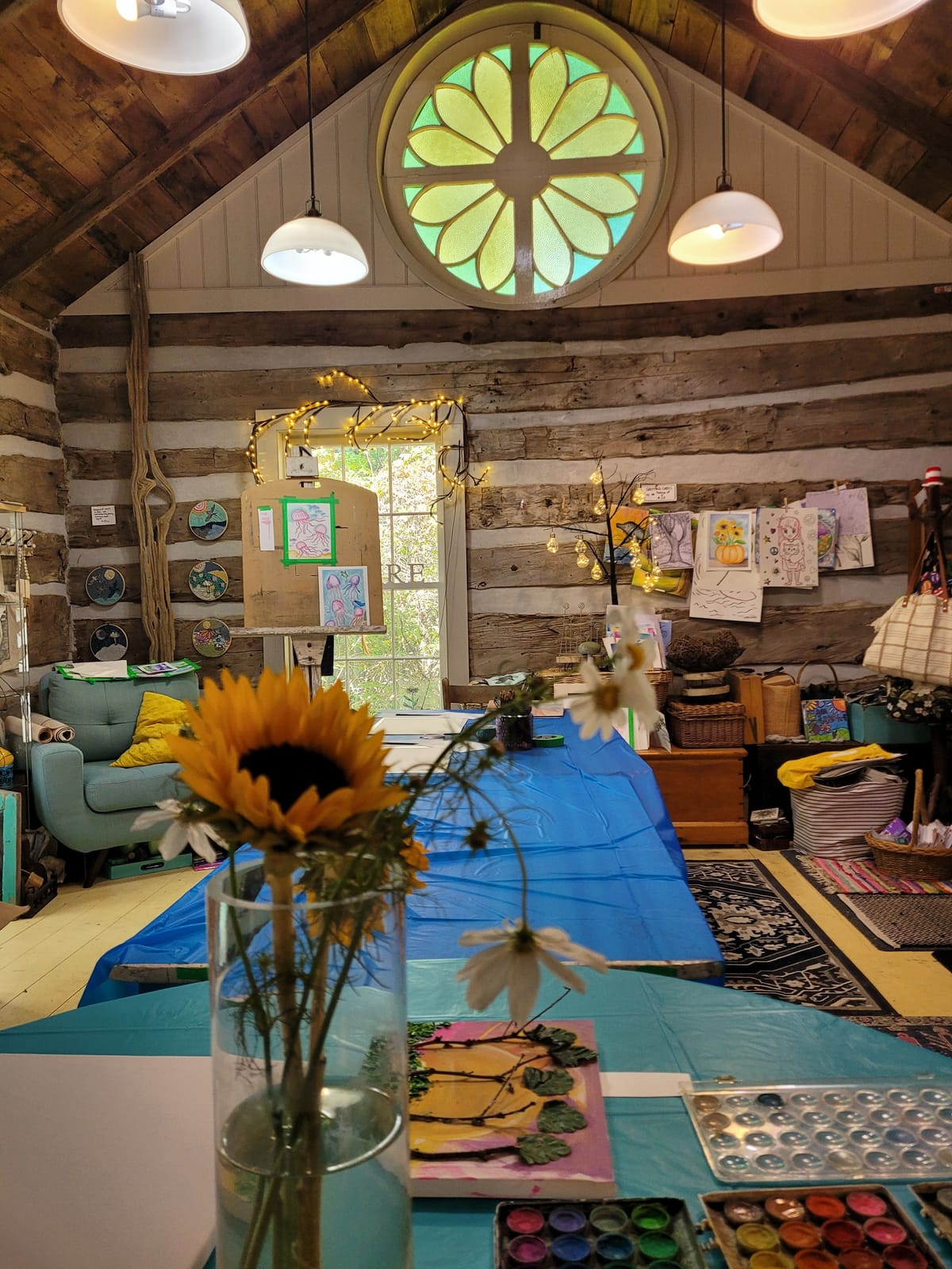 Inside the log-cabin studio: the round stained-glass window above an easel, a long table set for a class, and children's artwork on the walls.