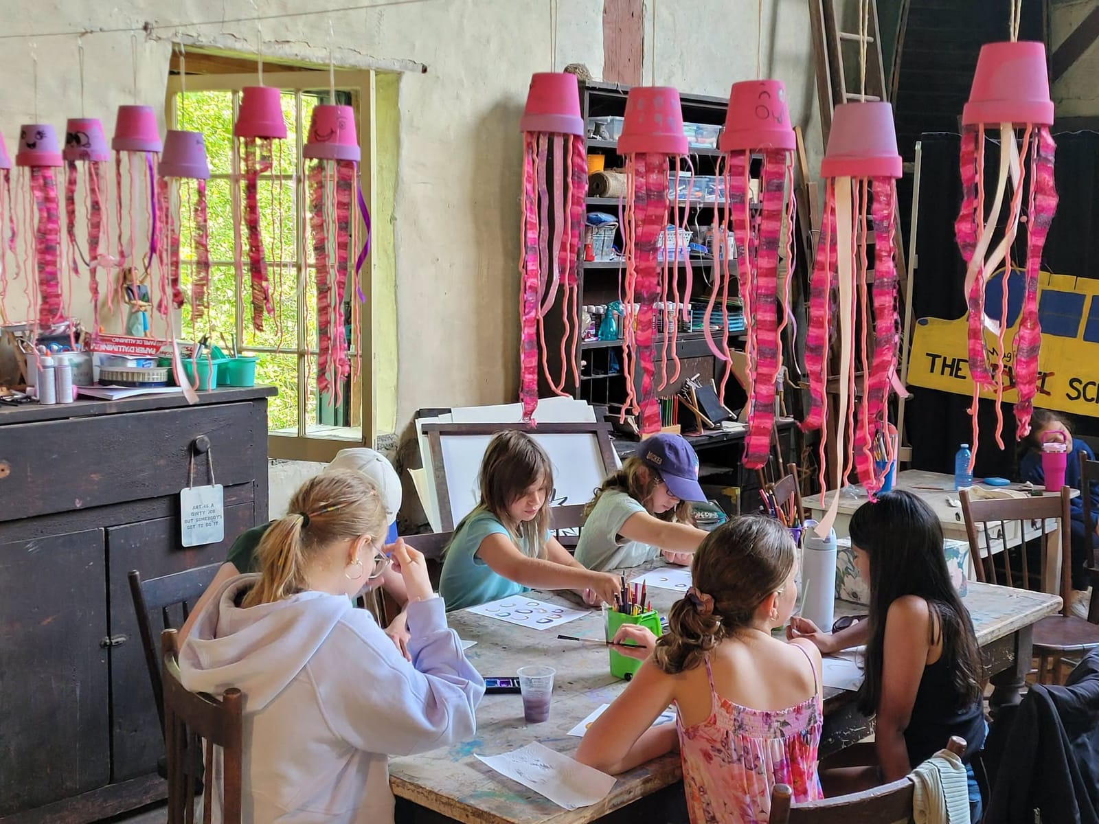 Girls drawing at a long table under hanging pink paper jellyfish.