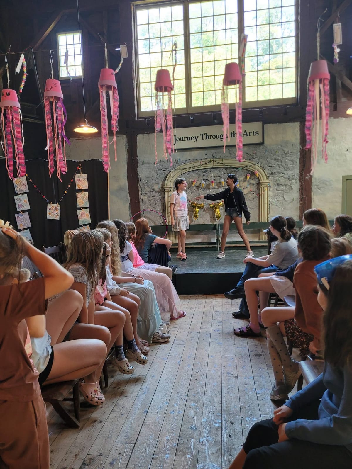 An audience watching children perform on a stage hung with pink paper jellyfish, under the Journey Through the Arts sign.