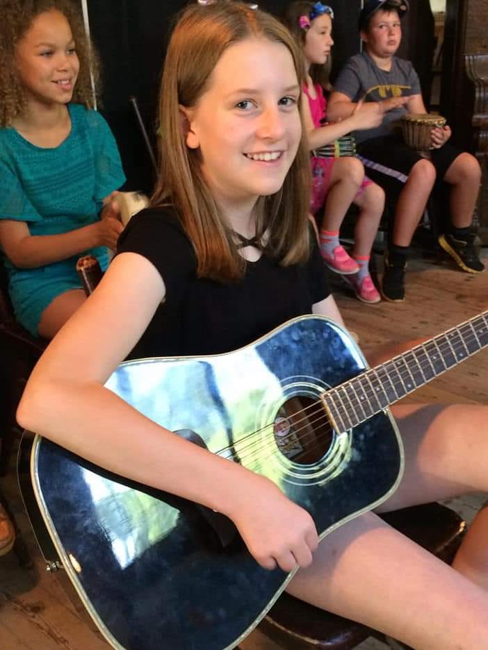 A smiling girl holding an acoustic guitar, with other campers seated behind her.