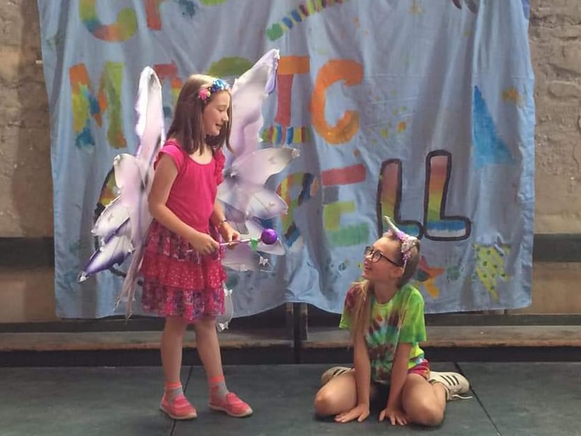 A girl in fairy wings performing on stage in front of a hand-painted banner, with another girl sitting on the floor beside her.