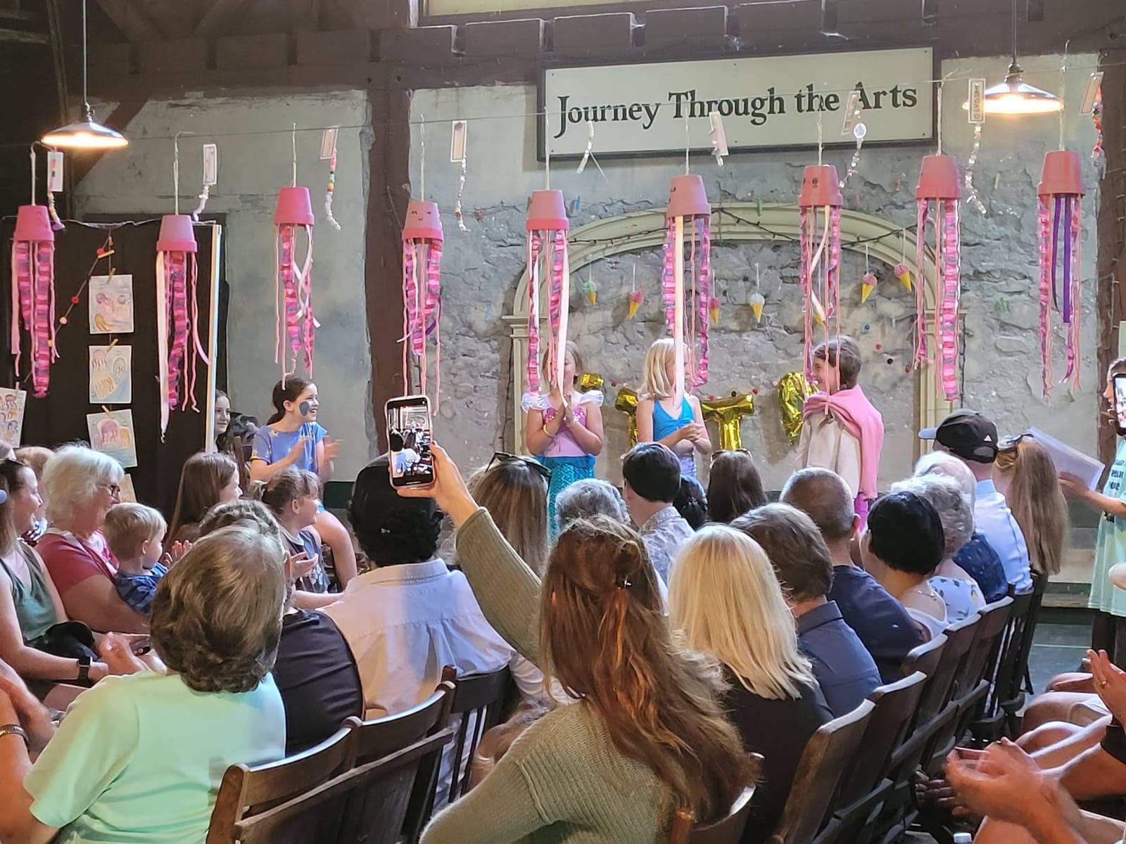 Families in rows watching campers perform on a stage decorated with pink paper jellyfish, under the Journey Through the Arts sign.