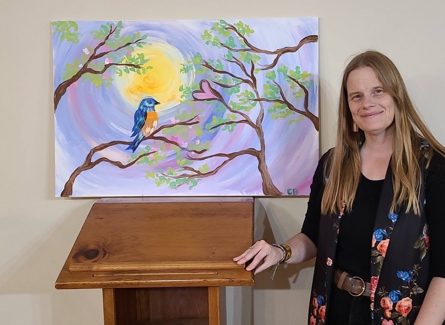 A woman standing beside an acrylic painting of a bluebird on a branch.