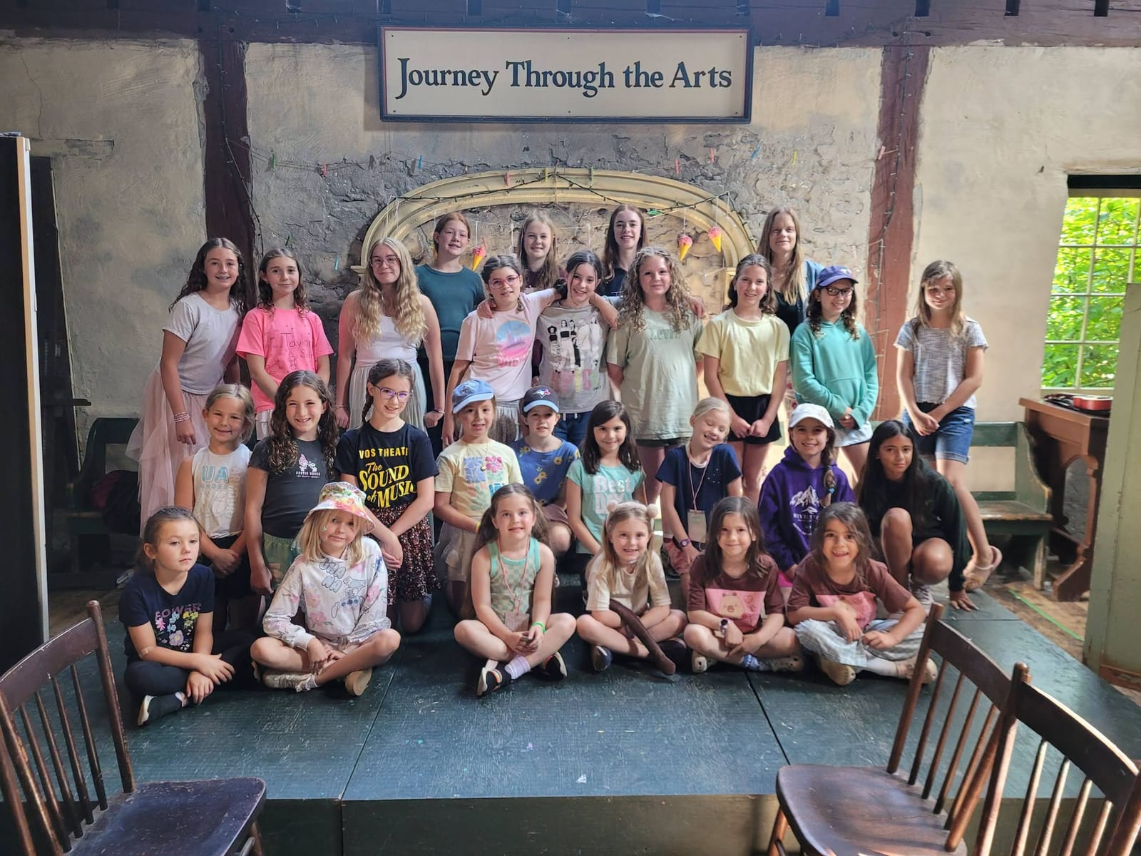A big group of campers posing together on the stage under the Journey Through the Arts sign.