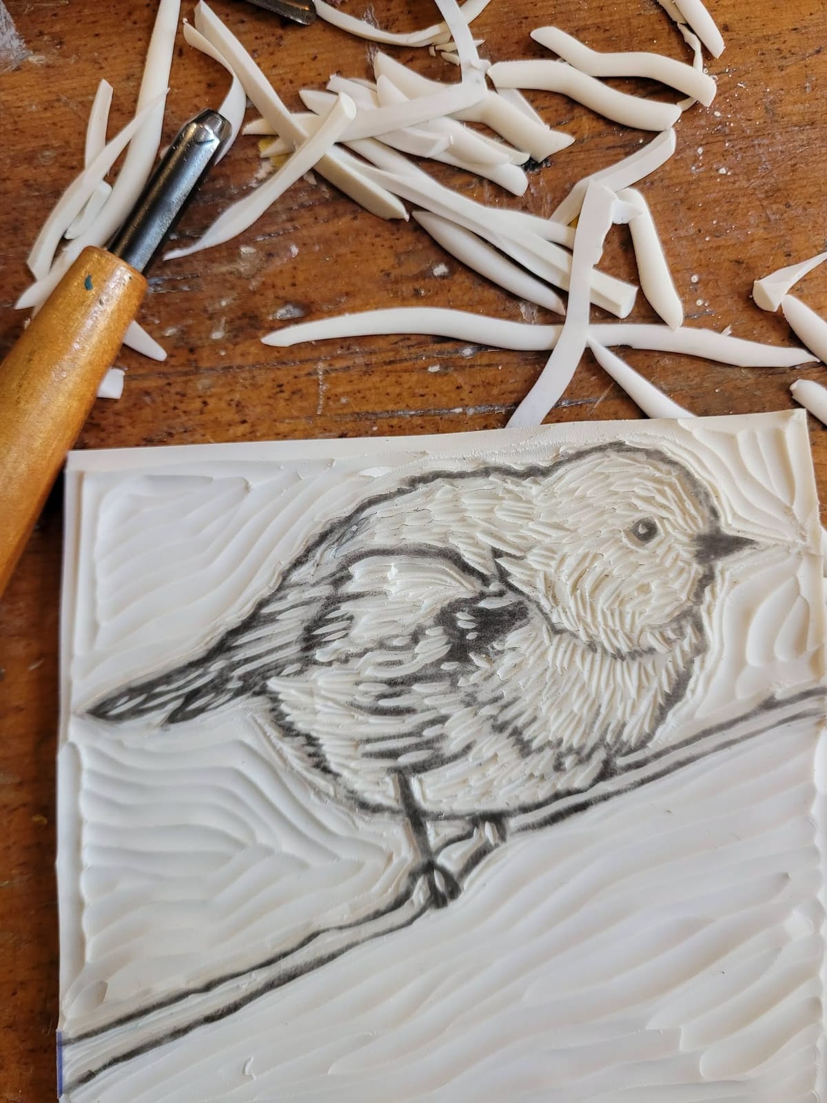 A carved lino block of a bird beside a carving tool and curls of cut lino.